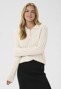 Wollmischpullover in Creme mit geripptem Muster, geknöpften Kragen und langen Ärmeln; kombiniert mit einem schwarzen, eng anliegenden Rock.