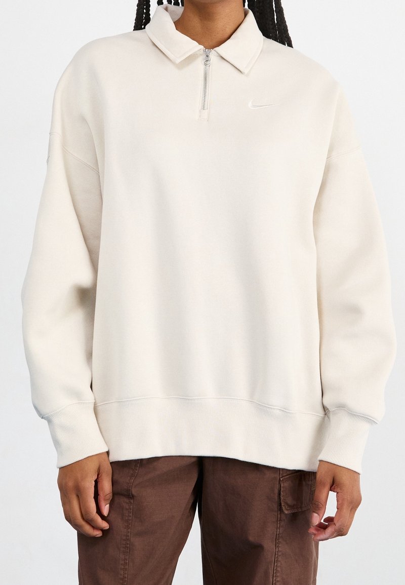 Sweatshirt zippé beige clair avec un col, des poignets et un ourlet côtelés. Présente un petit logo blanc sur la poitrine. Tissu doux et texturé.