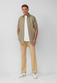 Hellgrünes kurzärmliges Button-Up-Hemd über einem weißen T-Shirt, kombiniert mit beigen Hosen und cremefarbenen Turnschuhen, vor einem schlichten Hintergrund.