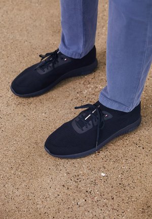 Personne portant des baskets noires en tricot avec des lacets et un pantalon bleu, debout sur un sol en béton texturé.