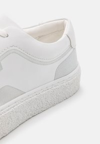 Zapatilla blanca con parte superior de cuero liso, acentos texturizados en gris y una suela de goma cremosa con manchas. Presenta costuras sutiles a lo largo del diseño.