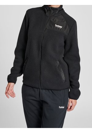 Osoba ubrana w czarną polarową kurtkę na zamek i czarne spodnie, oba z logo "hummel sportswear" na piersi i udzie.