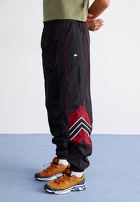 Pantalons de jogging noirs avec des accents rouges et des motifs en chevron ; fabriqués en matériau léger. Associés à des chaussures de sport orange et grises.