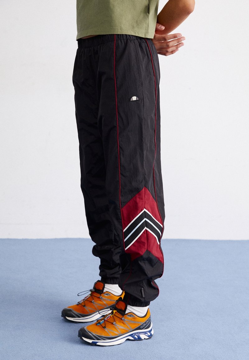 Pantalons de jogging noirs avec des accents rouges et des motifs en chevron ; fabriqués en matériau léger. Associés à des chaussures de sport orange et grises.