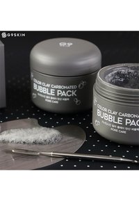 Des pots gris étiquetés "Color Clay Carbonated Bubble Pack", avec une texture en mousse sur un applicateur en acier inoxydable en forme de cœur, sur un fond à pois.