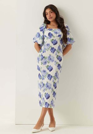 Donna con vestito midi bianco con stampa floreale blu e maniche a sbuffo, in piedi contro una parete liscia, con tacchi bianchi a punta.