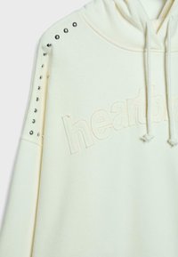 Sweat à capuche crème avec un logo "battement de cœur" texturé, des clous argentés sur les épaules et des cordons de serrage à la capuche. Tissu doux avec une coupe décontractée.