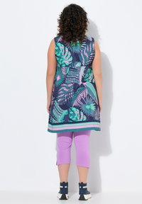 Bezramienna tunika na ciemnoniebieskim tle, z żywymi zielonymi i różowymi wzorami liści, w połączeniu z lawendowymi legginsami w 3/4 oraz sportowym obuwiem w kolorze granatowym.