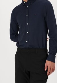 Námořnická modrá košile na knoflíky s herringbone vzorem, dlouhými rukávy a bílými knoflíky, spárovaná s černými kalhotami.