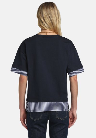 Blonde person wearing a navy short-sleeve top layered over a blue and white striped shirt, paired with dark blue jeans, shown from the back.