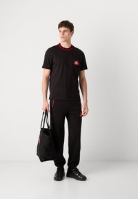 Zwarte katoenen t-shirt met rode kraag, met een klein geborduurd ontwerp. Gecombineerd met zwarte joggingsbroek met rode accenten. Man met een zwarte totebag.