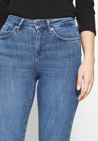 Vaqueros azules de mezclilla con un corte ajustado, cintura alta, bolsillos frontales, ligero desgaste y textura ligeramente deslavada. Cierre delantero con botones.