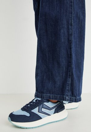 Navy and light blue athletic sneakers feature a textured suede upper, rounded toe, and white sole with light blue accents. Worn with dark denim.