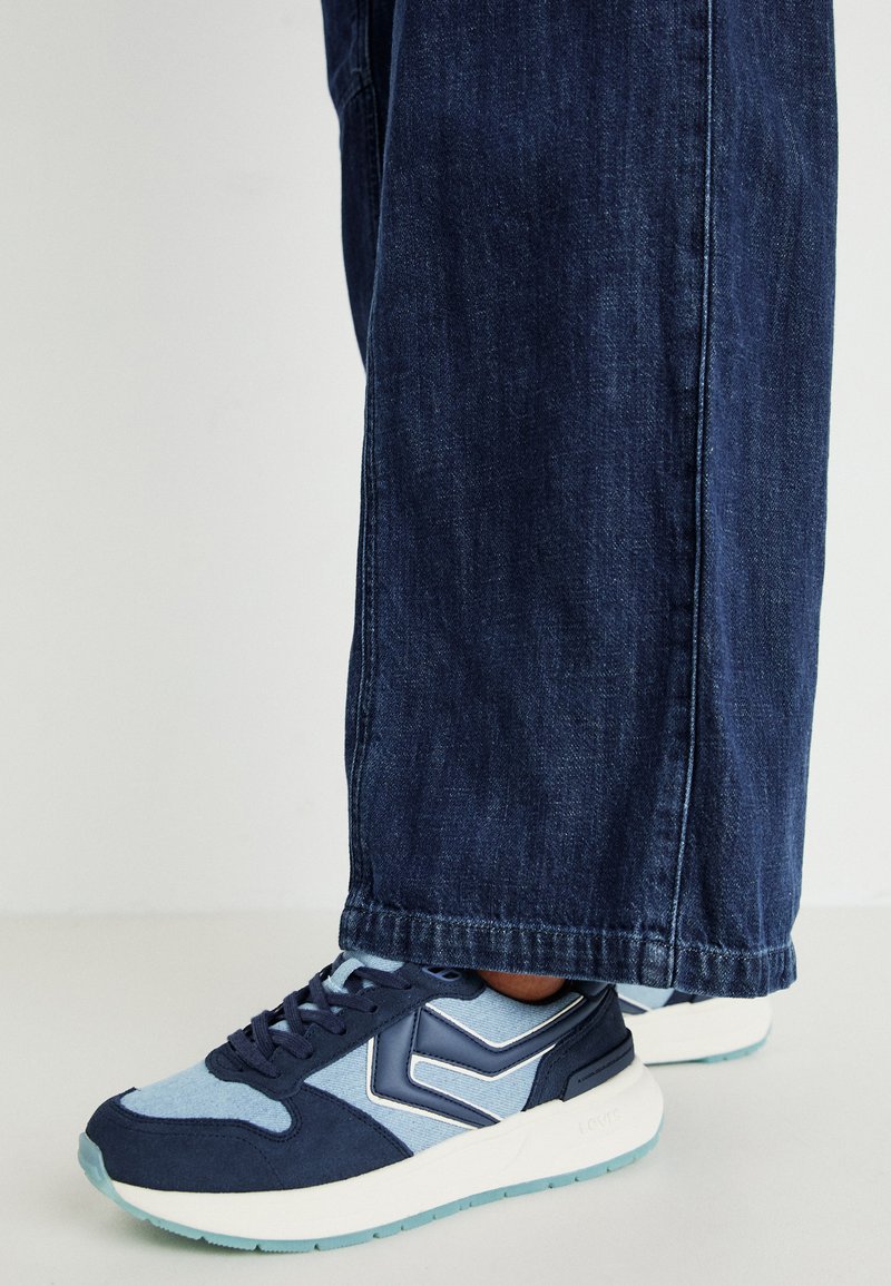 Tênis atléticos em azul marinho e azul claro apresentam um cabedal em camurça texturizada, bico arredondado e sola branca com detalhes em azul claro. Usados com jeans escuros.