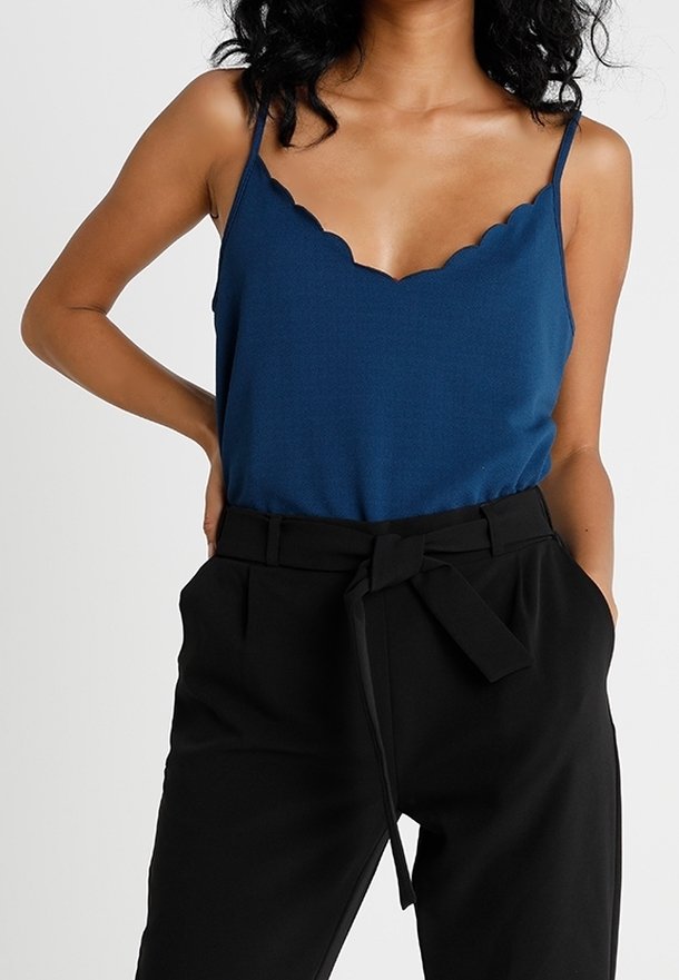 Femme portant un haut sans manches bleu à bords festonnés, rentré dans un pantalon noir taille haute avec ceinture nouée, mains dans les poches.