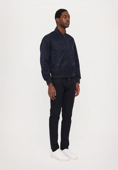 Navy bomber jacket with a smooth, shiny finish, ribbed cuffs, and collar. Worn with black pants and white sneakers.