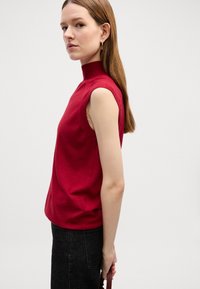 Top rosso senza maniche con colletto alto, realizzato in tessuto a maglia. Presenta una vestibilità rilassata e una texture liscia. Indossato con jeans scuri.