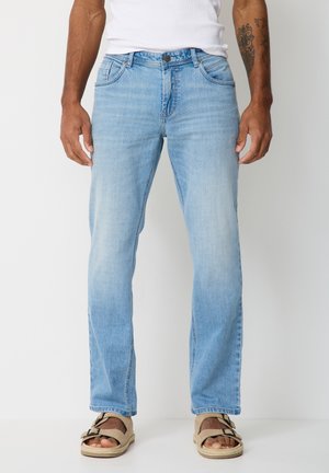 Hellblaue Jeans mit geradem Bein, getragen von einer Person mit gebräunter Haut, kombiniert mit beigen Sandalen und einem weißen ärmellosen Oberteil.