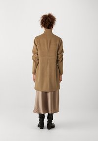Cappotto di lana beige con colletto strutturato, vestibilità rilassata e dettagli di cucitura, abbinato a una gonna di raso e stivali neri fin sopra il ginocchio.