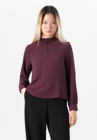 Kvinne med rett, lys ombre hår iført en langermet mørk lilla bluse med volangkrage og svarte bukser, stående mot hvit bakgrunn.