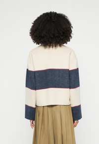 Κομμένο μπουφάν σε κρεμ και τζιν μπλε με κόκκινες ρίγες ως τονισμός. Χαλαρή γραμμή, φαρδιά μανίκια, ομαλή υφή. Συνδυασμένο με πλισέ φούστα σε ταμπά απόχρωση.