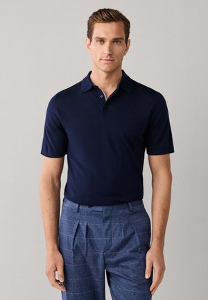 Man wearing a navy short-sleeve polo shirt and blue checked pleated trousers standing against a plain light background.