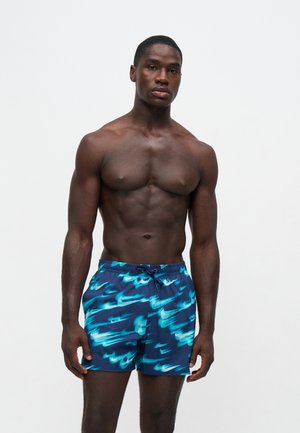 Uomo senza maglietta con corporatura muscolosa che indossa pantaloncini da bagno blu con un motivo Nike swoosh luminoso su uno sfondo bianco semplice.