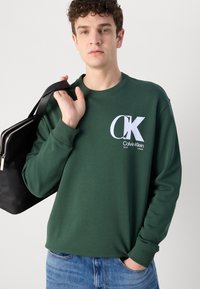 Pulover verde cu mansete și guler ribate, având un logo alb Calvin Klein pe piept. Modelul ține o geantă neagră.