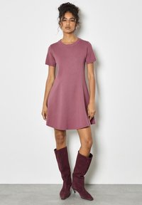 Abito a maniche corte a forma A in tessuto malva, con scollatura rotonda e dettagli sulle cuciture. Abbinato a stivali al ginocchio in suede bordeaux.