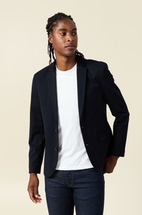 Marinblå kavaj med en åtsittande design, enkel knappstängning och slagkrage. Lättviktig tyg, sidofickor, kombinerad med en vit t-shirt.