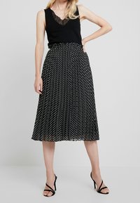 Černý top bez rukávů s krajkovým lemem, v kombinaci s černou sukní střední délky s bílými puntíky a skladovanou texturou. Černé lodičky.