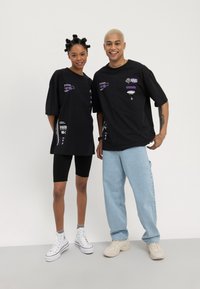 Black oversized t-shirts with graphic prints in purple, paired with black bike shorts and sneakers. Light blue jeans and beige shoes complete the look.
