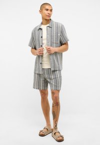 Pánský dvoudílný outfit obsahující krátké rukávy, pruhovanou košili s knoflíky v šedé a krémové barvě, odpovídající šortky a světle hnědé sandály.