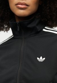 Gros plan d'une personne portant une veste noire zippée avec le logo blanc d'Adidas et trois bandes sur les épaules.
