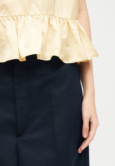 Gros plan d'une personne portant un haut jaune pâle à volants et un pantalon bleu marine foncé, avec le bras droit et la main détendus le long du corps.