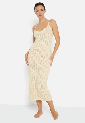 Woman with dark hair in a beige sleeveless knitted midi dress standing barefoot against a white background.