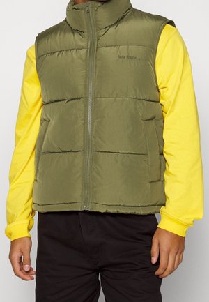 Man wearing olive green puffer vest with zipper over bright yellow long sleeve shirt and black pants, standing with arms relaxed at sides.