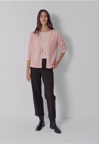 Cardigan côtelé rose pâle sur un haut rose clair, assorti à un pantalon noir taille haute et des bottines noires. Design simple, fermeture par boutons.