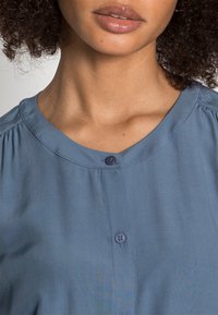 Blouse bleu clair avec un col rond, des plis subtils et une fermeture à un bouton à l'avant. Tissu lisse avec une légère brillance.