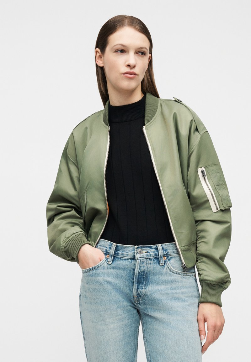 Jeune femme portant une veste bomber vert clair par-dessus un pull à col roulé noir et un jean bleu clair, debout avec une main dans la poche.