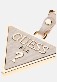 Portachiavi a forma di triangolo in materiale beige testurizzato con dettagli dorati; presenta la scritta "GUESS USA" in rilievo e un punto interrogativo decorativo.