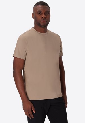 Uomo con capelli corti che indossa una T-shirt beige a maniche corte e pantaloni neri, in piedi davanti a uno sfondo chiaro e uniforme.