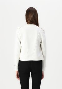 White cropped jacket with a structured design, featuring a pointed collar and back seams. Sleeves have black detailing. Worn with black pants.