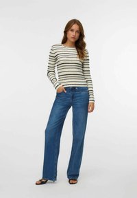 Donna che indossa un maglione a maniche lunghe bianco e nero a righe, jeans larghi blu e tacchi aperti neri, in piedi con una mano in tasca.