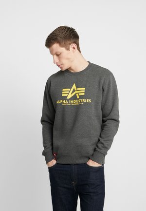 Grauer Sweatshirt aus Baumwollmischung; verfügt über ein gelbes Logo und Text auf der Vorderseite, Rundhalsausschnitt und gerippte Bündchen. Kombiniert mit dunklen Jeans.