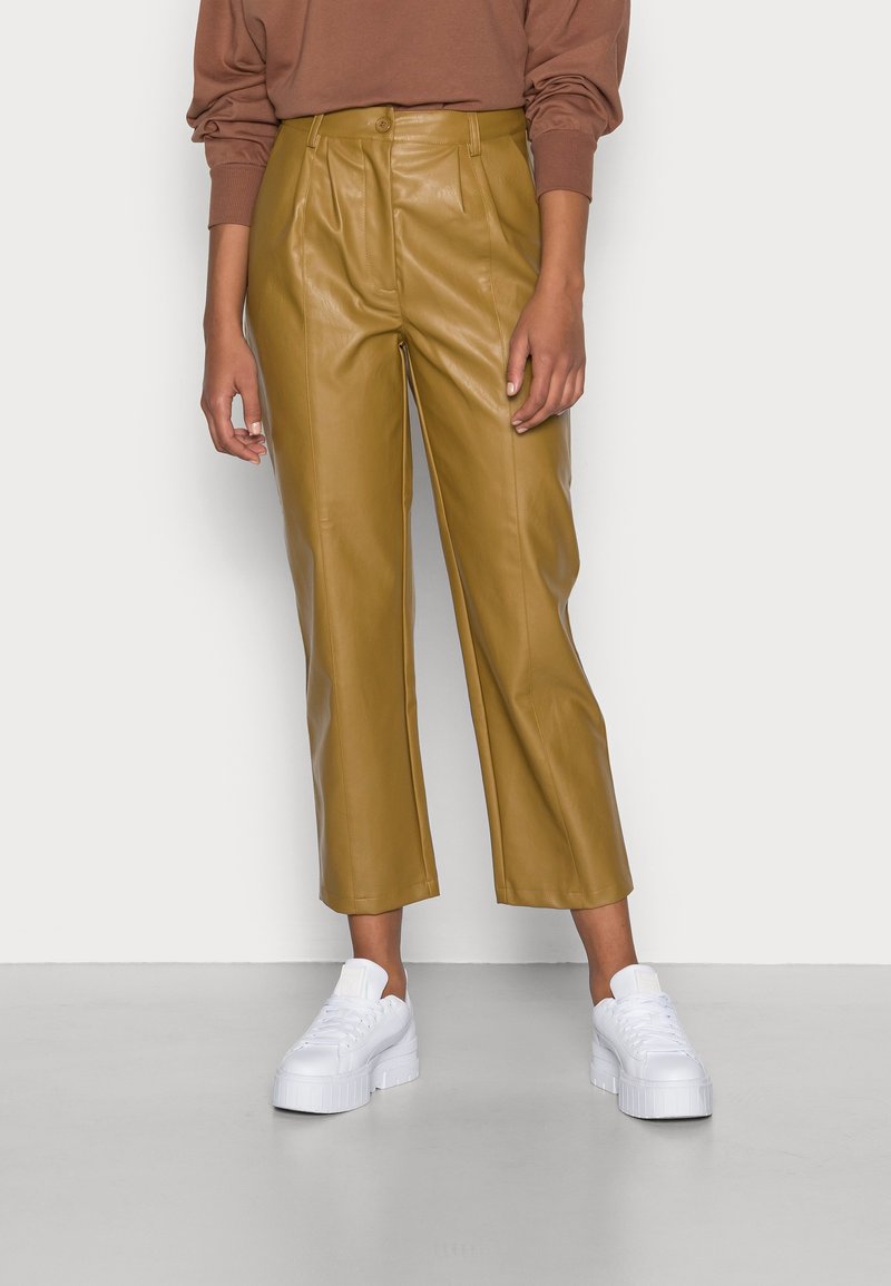 Femme portant un pantalon en cuir court couleur moutarde, un haut marron à manches longues et des baskets blanches à plateforme, debout contre un mur blanc.