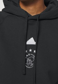 Haut à capuche noir avec un cordon de serrage, logo Adidas blanc et design graphique avec des étoiles et un motif de personnage sur le devant.