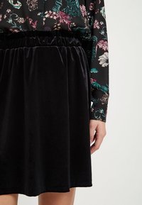 Jupe en velours noir avec une taille élastique, présentant une texture lisse et une coupe longueur genou, associée à un chemisier à motifs floraux.