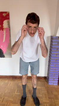 Άνδρας με μουστάκι που προσαρμόζει τα γυαλιά του, φοράει λευκό μπλουζάκι, τζιν σορτς, γκρι κάλτσες και μαύρα παπούτσια, στέκεται μέσα σε εσωτερικό χώρο δίπλα σε έργο τέχνης και μοβ τούβλινο τοίχο.