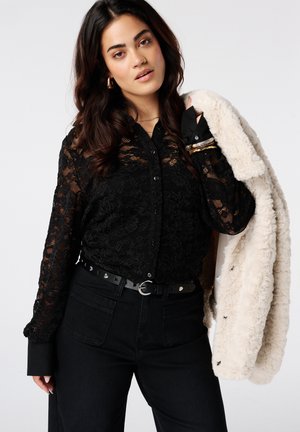 Femme aux longs cheveux foncés portant un chemisier en dentelle noire et un pantalon noir, tenant un manteau en fausse fourrure blanche sur l'épaule, regardant la caméra.